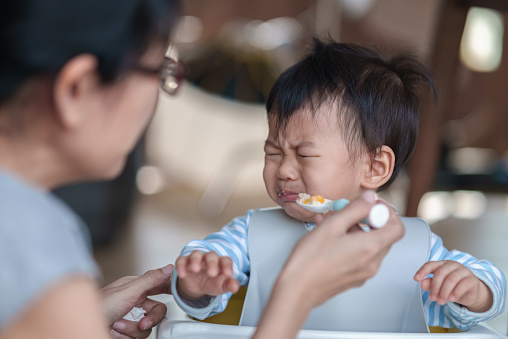 Asian baby boy baby boy refusing to eat food and crying while his mother feeds him. Mother try to feeding her son with spoon. Children daily lifestyle take care or toddler nutrition food prepare concept