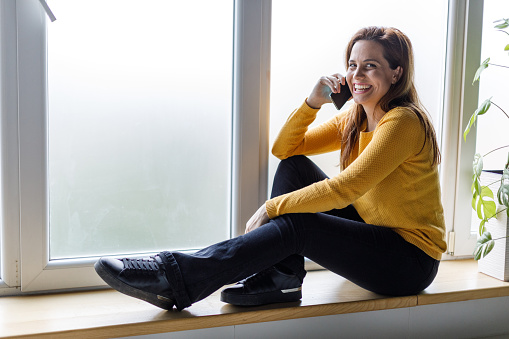 Full length shot of happy young woman sitting on the window sill, talking on the phone with a friend, looking at camera and smiling joyfully.