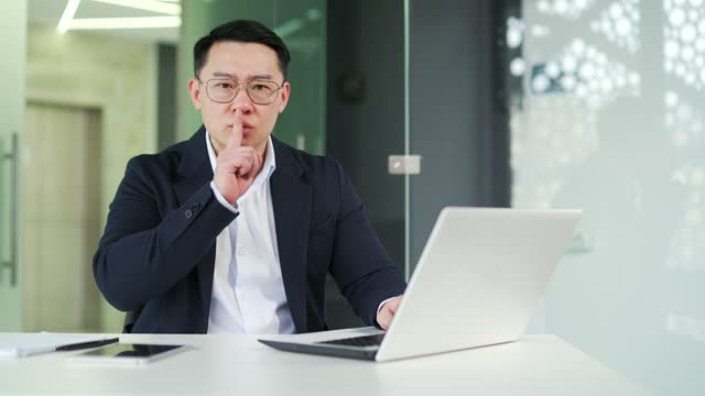 Asian man holding index finger to lips while working on laptop while sitting at workplace in office.