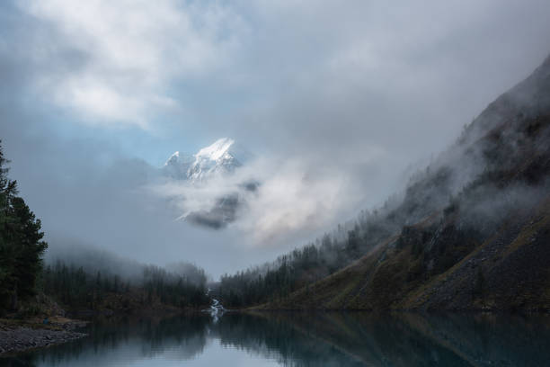 istock Tranquil scenery with snow castle in clouds. Mountain creek flows from forest hills into glacial lake. Snowy mountains in fog clearance. Small river and coniferous trees reflected in calm alpine lake. 1488123813
