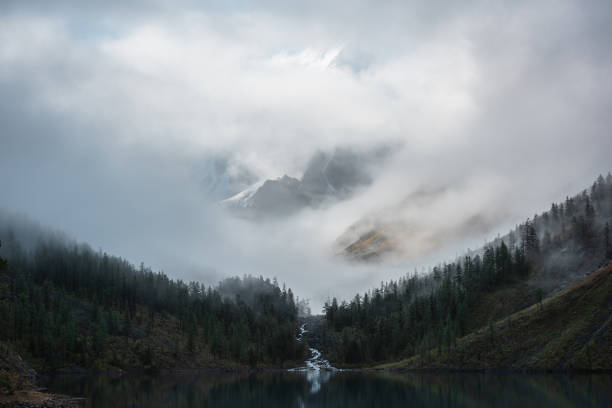 istock Tranquil scenery with snow castle in clouds and alpine lake. Mountain creek flows from forest hills into glacial lake. Snowy mountains in fog clearance. Small river and coniferous trees in low clouds. 1488123835
