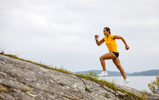 Side view of fitness woman running high-intensity interval training on a steep hill up a mountain by the sea. HILIT workout.