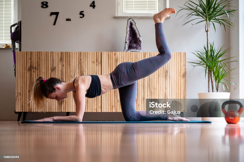 Fit young woman in sportswear working out at home in living room Fit young woman in sportswear working out at home in living room on a mat. Making a back kick exercise for toned glutes and legs. 35-39 Years Stock Photo