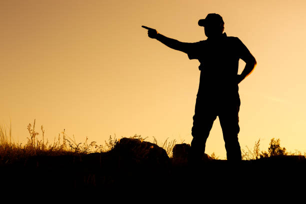 the silhouette of a man in a cap points with his hand to the left direction or something. outdoor grass and stones
