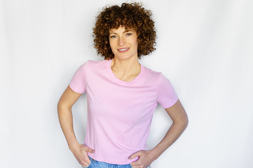 Portrait of emotionless adult curly haired female in pink t-shirt looking at camera against white background
