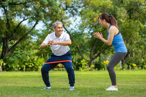 Senior asian man is using rubber band to build up his leg muscle strength while his daughter is cheering up in the public park for elder longevity exercise and outdoor workout