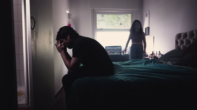 Couple in crisis: Young man sitting by bedside covering face in frustration while girlfriend stands in background feeling disconnected in the relationship