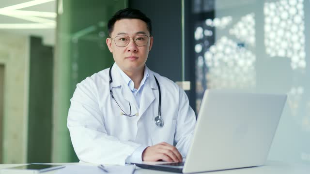 Portrait of a serious asian doctor in a white coat using a laptop while sitting at a workplace in an office in a hospital clinic.