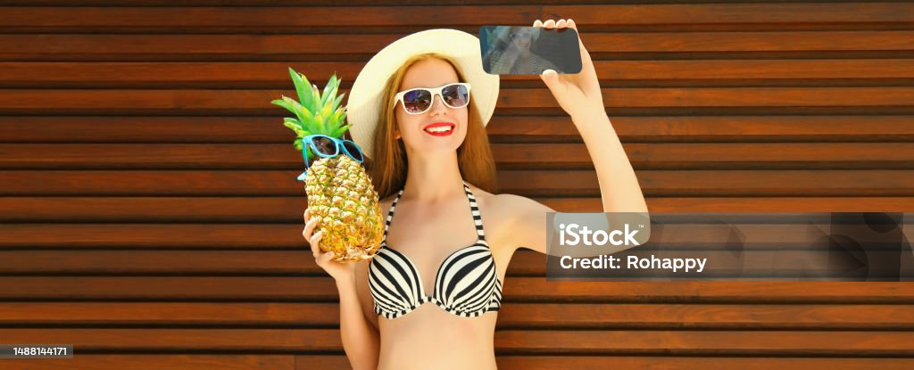 Summer portrait of happy smiling woman holding pineapple taking selfie with smartphone wearing straw hat, sunglasses, bra 25-29 Years Stock Photo