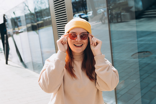 Happy cheerful young woman walking on city street. Portrait of beautiful woman in yellow hat and pink sunglasses smiling outdoors. Urban lifestyle concept.