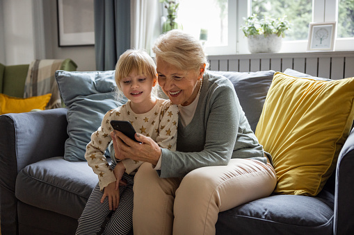 Senior woman sitting with small girl in living room at home using smart phone  for video call or playing games.