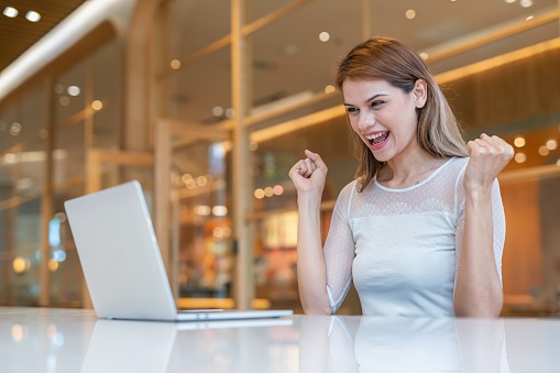 Close up mid shot of multiracial businesswoman using laptop cheering for success from a workspace with cafe working out of home, from a workspace shared office cafe.