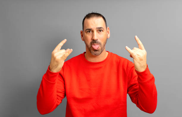 латиноамериканец с бородой в возрасте 40 лет, одетый в красный свитер, делает рок-жест, высунув язык в сумасшедшей позе, изолированный на жел� - making a face mischief sticking out tongue isolated on red стоковые фото и изображения