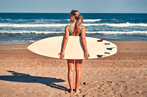Beautiful caucasian woman in a swimsuit with a surfboard standing on a sandy beach near the sea. Sports and active recreation.