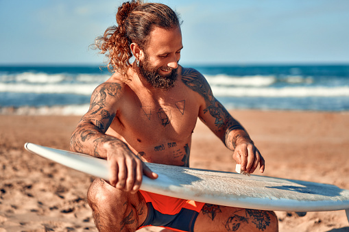 Caucasian beefy bearded man with tattoos in swimming trunks is waxing a surfboard near the sea. Sports and active recreation.