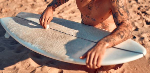 Caucasian beefy bearded man with tattoos in swimming trunks is waxing a surfboard near the sea. Sports and active recreation.