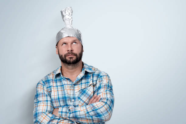 Funny bearded man in a plaid shirt with an aluminum hat looks up, standing next to a light wall background. Funny bearded man in a plaid shirt with an aluminum hat looks up, standing next to a light wall background. tin foil hat stock pictures, royalty-free photos & images