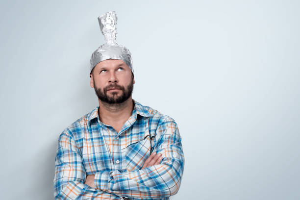 funny bearded man in a plaid shirt with an aluminum hat looks up, standing next to a light wall background. - tin foil hat imagens e fotografias de stock