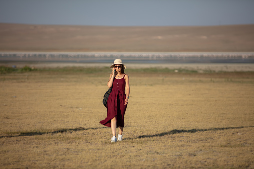 A blonde woman in a red dress is walking on the steppe. She is talking on mobile phone.
