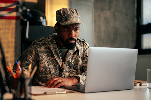 A US army soldier is in his uniform in the office, working on his laptop.