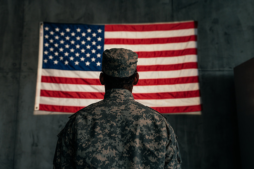 An American soldier is standing in front of an American flag on the wall, shot from the rear.