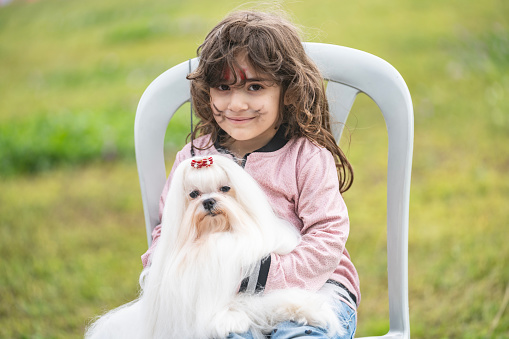 Portrait of little girl holding long white haired Maltese pet dog in outdoor. They are sitting in public park. Shot under daylight.