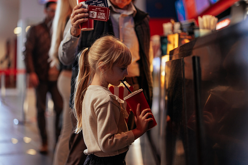 A small Caucasian child is drinking soda while waiting for her parents to pay for the movie tickets in the theater.