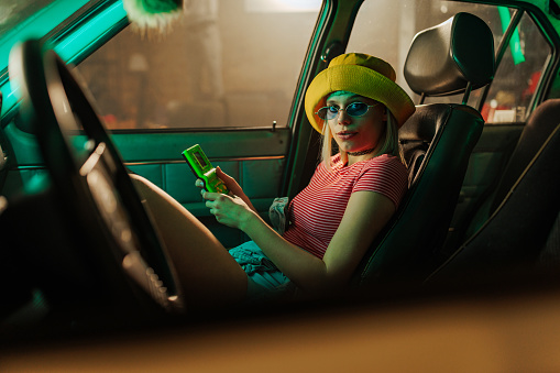 A retro styled woman is sitting on the passenger seat in the car holding a vintage video game in her hands and looking at the camera.