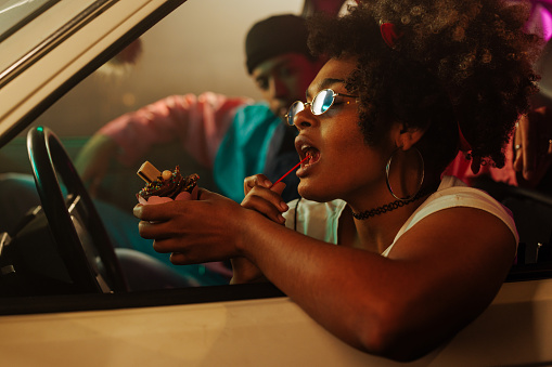 A fashionable retro girl is sitting in the car eating an ice cream while her boyfriend is sitting in the passenger seat.