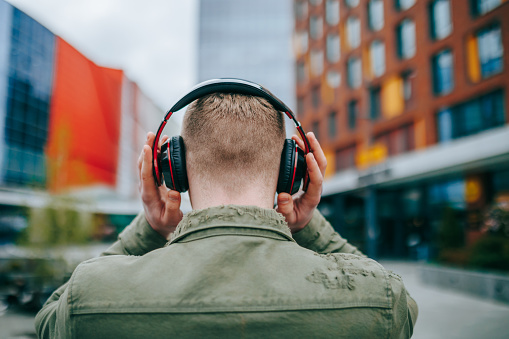 A handsome caucasian man listening to music with his headphones on and looking at the beautiful city in the background, seen from the back.