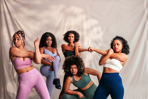 A group of confident diverse skin toned athletic women are stretching indoors in their activewear.