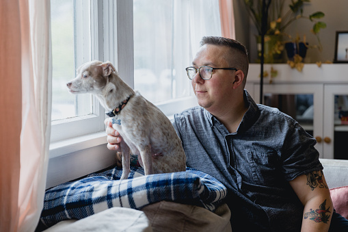 Portrait of an autistic man with his dog indoors. They are sitting on their couch in the living room of his home. Shot using natural window light.