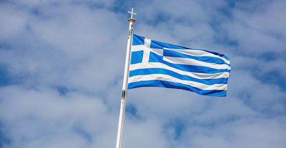 Greece sign symbol. Greek national official flag in blue and white on flagpole waving in wind. Cloudy sky background, sunny day in Cyclades island.