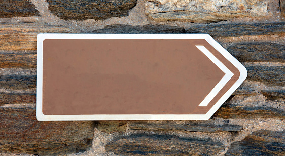 Metal directional signboard empty blank, brown signpost with white arrow put on rocky wall construction background. Cyclades, Greece. Copy space