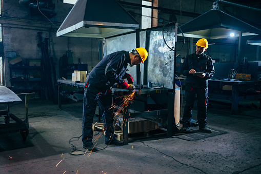 A supervisor is in the factory workshop observing a senior Caucasian worker grinding metallic parts.