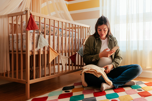 A young adult smiling pregnant woman is at home sitting in the nursery using her cellphone.