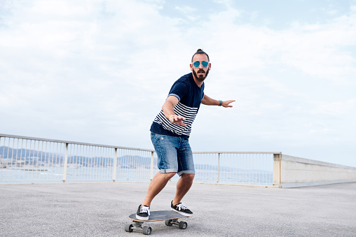 Full length of cool bearded hipster in sunglasses balancing on wide longboard while riding and having fun on paved waterfront