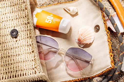 Sunscreen and sunglasses in a woven straw bag on a sandy beach