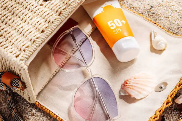 Photo of Sunscreen and glasses in a wicker straw bag on a sandy beach