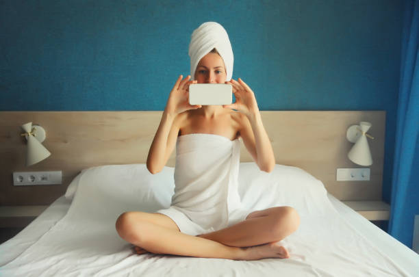 mulher jovem feliz tirando selfie com telefone secando seu cabelo molhado após o banho com toalha enrolada em sua cabeça sentada na cama pela manhã - hotel domestic room telephone technology - fotografias e filmes do acervo