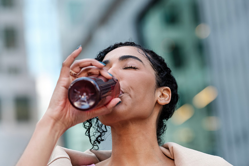 Headshot of young fitness woman drinking energy drink from a bottle outdoors. Blurry background and her eyes are closed. She looks relaxed. Resting and hydrating in sports concept.