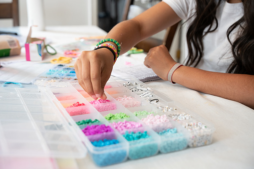 A girl sits at her kitchen table making bracelets for a fundraiser, carefully stringing beads and tying knots with a smile on her face, hoping to make a positive impact.