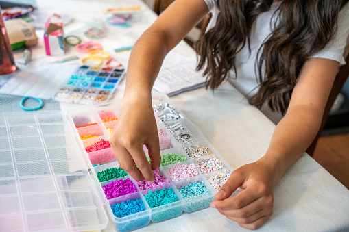 A girl sits at her kitchen table making bracelets for a fundraiser, carefully stringing beads and tying knots with a smile on her face, hoping to make a positive impact.