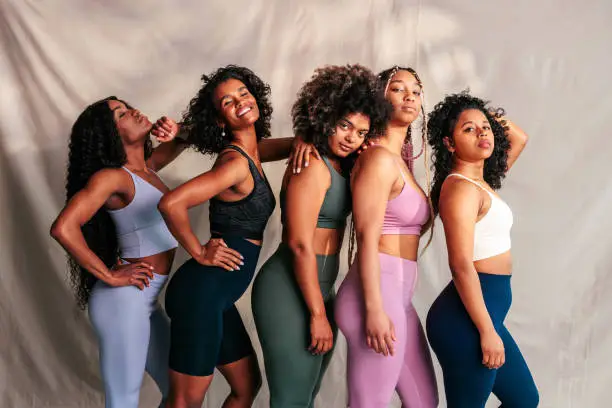 A group of young trendy athletic women are indoors, posing together in a line and looking at the camera.