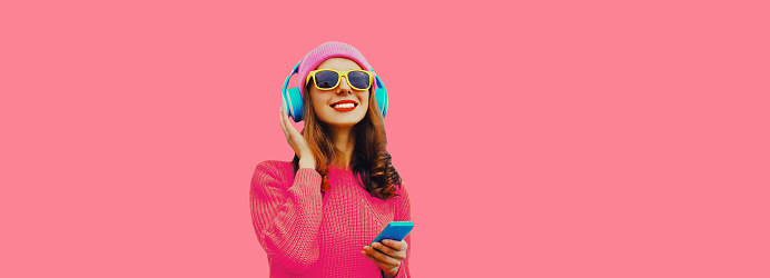 Portrait of happy smiling modern young woman in wireless headphones listening to music with smartphone wearing knitted sweater, hat on pink background