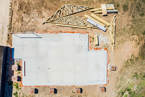 Aerial view looking directly down on house construction site: reinforced concrete waffle slab foundation or floor has been laid; girder frame for garage entrance in place; stacks of bricks for the walls are in position; timber roof trusses, rafters and battens on the ground with labelled timber noggins for strengthening.