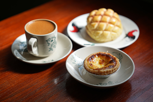 A freshly baked Hong Kong style polo bun and egg tart, served with a cup of milk coffee, on a wooden table in a quaint local tea shop