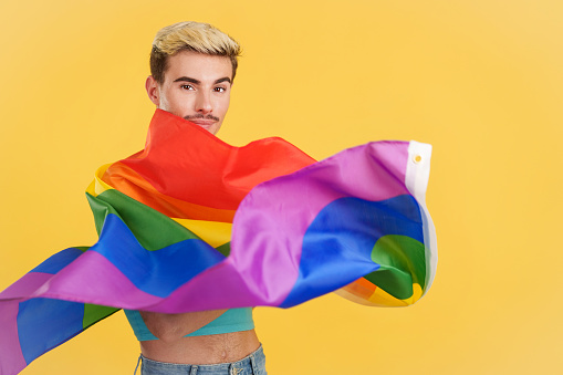 Happy gay man waving a lgbt rainbow flag in studio with yellow background