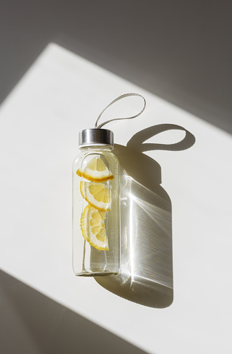 Lemon water drink detox in bottle, hard shadow at sunlight on white background. Wellness, diet, eating healthy concept. Stylish glass reusable water bottle, eco friendly lifestyle minimal trend photo