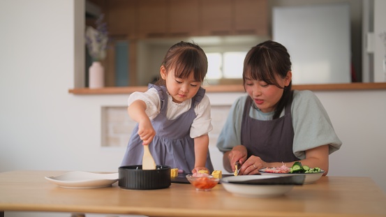 A mother and her daughter is preparing sushi together at home. The mother is assisting her small daughter making sushi together.